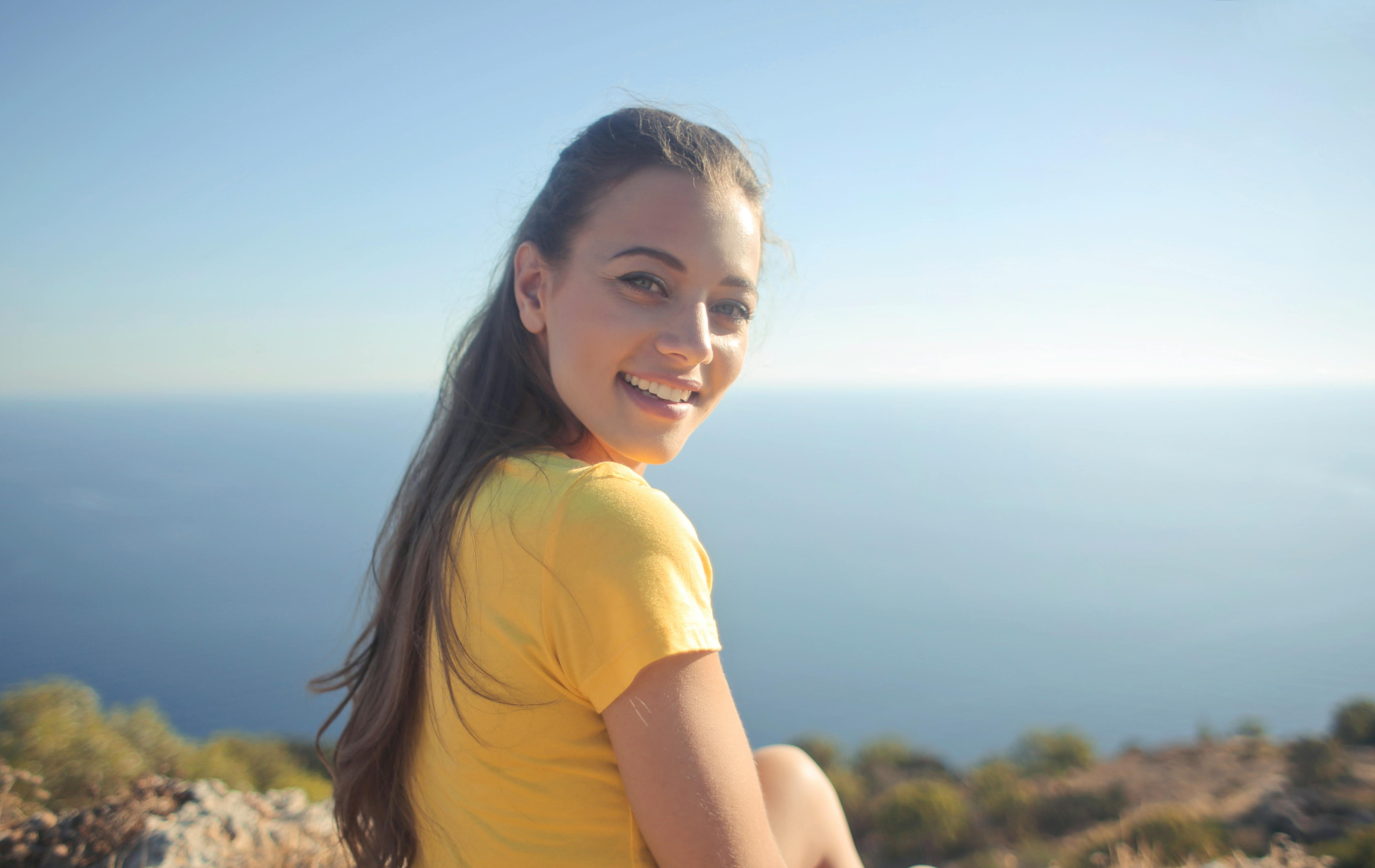 Young woman in a yellow shirt smiles by the sea, enjoying the outdoor view.